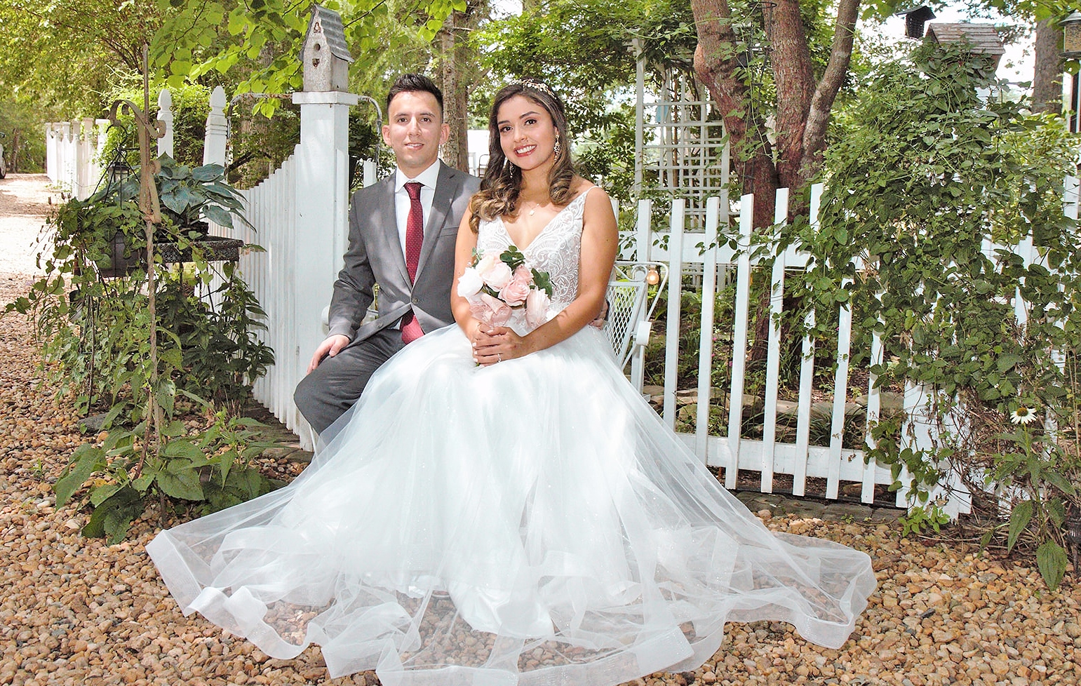 Bride and groom pose on white bench in front of picket fence.