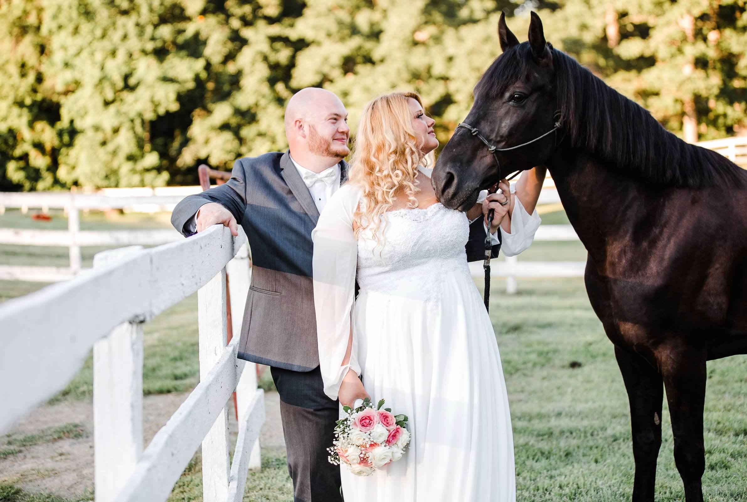 Bride pets black horse while groom leans against the white board fence.