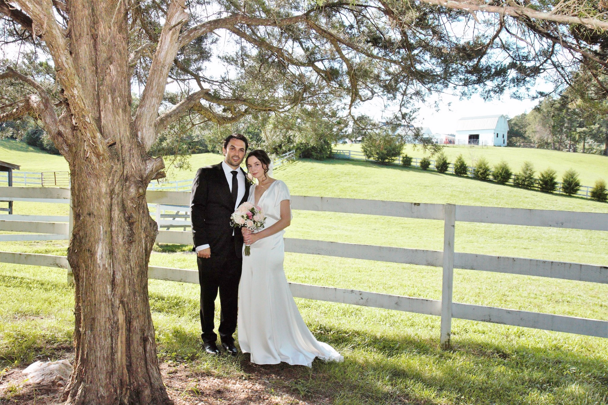 Bride and groom posing next to old cedar tree in front of pasture.