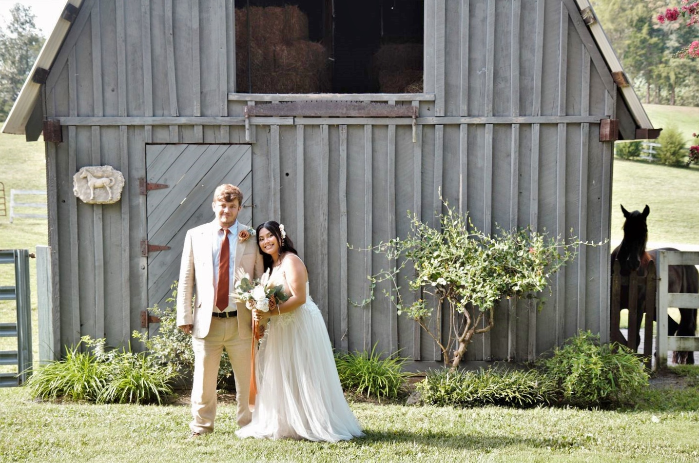 Bride and groom and black horse pose in front of the gray barn.