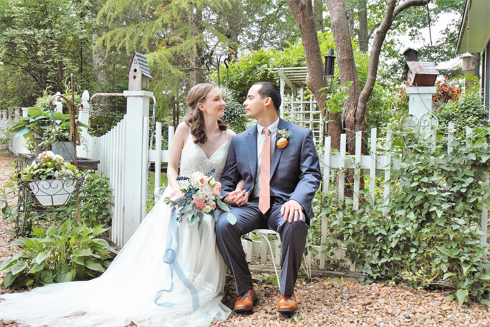 Bride and groom sitting on the white iron bench in front of the picket fence.